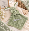 A light green plush bunny security blanket with the name 'Blake' embroidered in white script on the front, styled on a cream knit blanket with baby props.