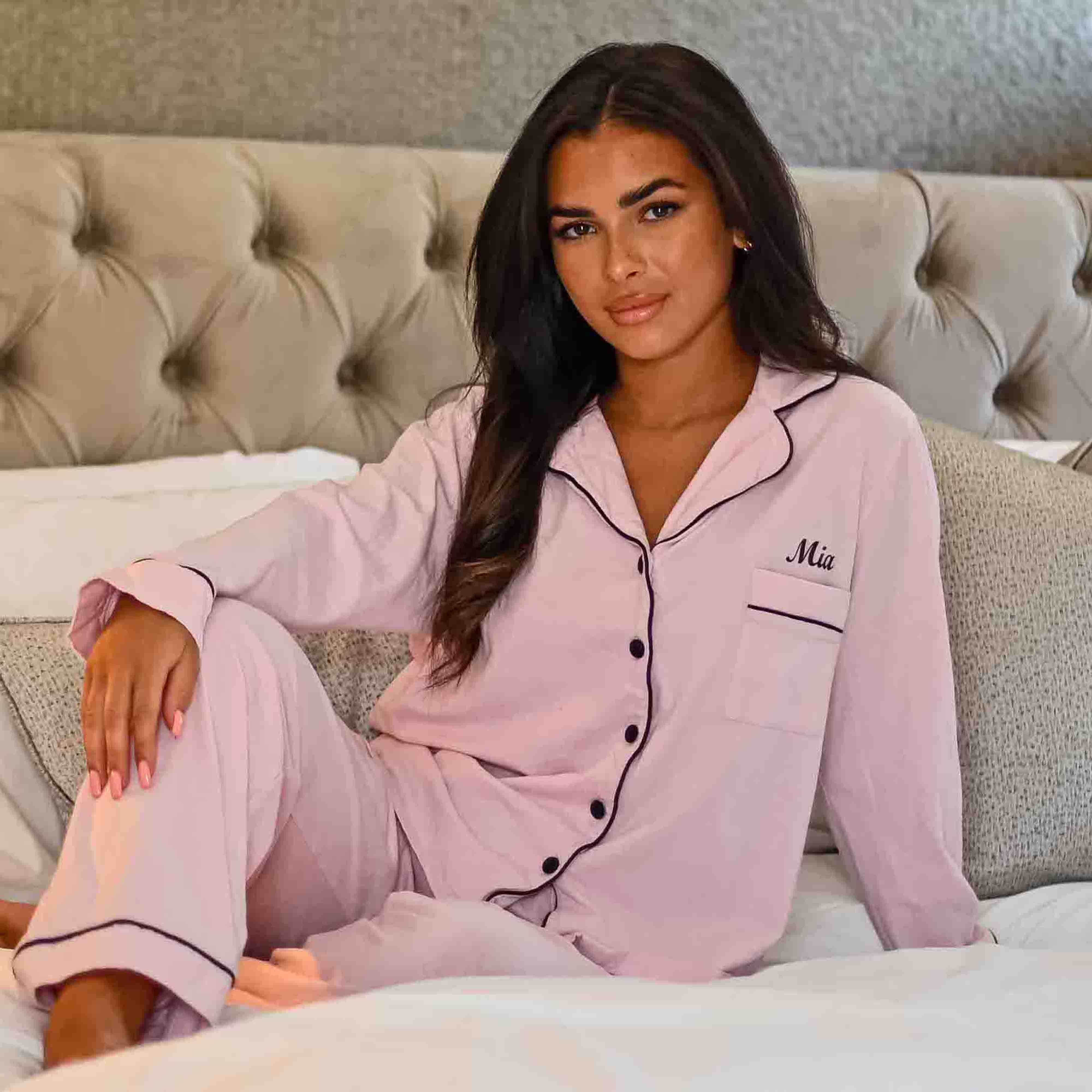 Lady wearing a Light Pink personalised pyjama set. Long length trousers with a satin trim to the bottom, tie around the waist and pockets on either side. Complemented with a long sleeve button up shirt with satin contract piping and buttons, a pocket to the right of the shirt pocket as you look at it. Completed with personalisation above the pocket. This can be personalised with whatever you want. The perfect personalised pyjama set for ladies. 