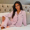 Lady wearing a Light Pink personalised pyjama set. Long length trousers with a satin trim to the bottom, tie around the waist and pockets on either side. Complemented with a long sleeve button up shirt with satin contract piping and buttons, a pocket to the right of the shirt pocket as you look at it. Completed with personalisation above the pocket. This can be personalised with whatever you want. The perfect personalised pyjama set for ladies. 