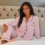 Load image into Gallery viewer, Lady wearing a Light Pink personalised pyjama set. Long length trousers with a satin trim to the bottom, tie around the waist and pockets on either side. Complemented with a long sleeve button up shirt with satin contract piping and buttons, a pocket to the right of the shirt pocket as you look at it. Completed with personalisation above the pocket. This can be personalised with whatever you want. The perfect personalised pyjama set for ladies. 

