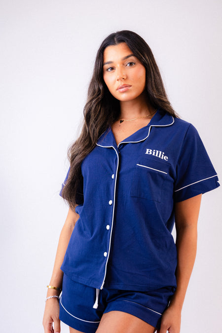 Ladies short jersey pyjamas in Navy with a White contrast satin piping and buttons. Short sleeve button shirt with a pocket to the right of the chest and your personalised name above. Shorts with both pockets and an adjustable tie around the waist. 