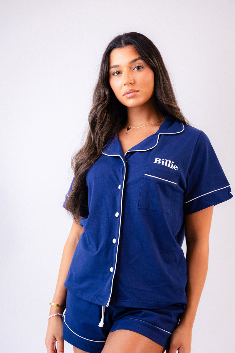 Ladies short jersey pyjamas in Navy with a White contrast satin piping and buttons. Short sleeve button shirt with a pocket to the right of the chest and your personalised name above. Shorts with both pockets and an adjustable tie around the waist. 