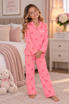 A young girl stands barefoot in a cozy bedroom wearing a pink pajama set with a bow print. The set includes a long-sleeve top and matching pants, with the name 'Amelia' embroidered on the chest.