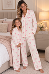 A woman and a child wearing matching pajama sets in a cozy bedroom. The pajamas are off-white with a pink bow print and pink piping, with the name 'Sophie' embroidered on the woman's collar.