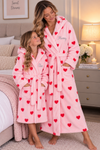 A woman and a child wearing matching pink hooded bathrobes with red heart prints, standing in a cozy bedroom setting. The woman's robe is personalized with the name 'Mummy' and the child's with 'Ellie' in gray script.