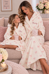 A woman and a young girl sitting together on a couch wearing matching plush bathrobes. The robes are cream-colored with a light pink bow pattern, and both have the name embroidered on the chest.