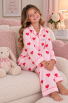 A young girl sits on a cozy pink couch wearing a light pink pajama set with red hearts and piping. The pajamas are personalized with the name 'Andrea' on the chest.