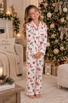 A young girl stands barefoot in a cozy living room wearing a matching pajama set with a festive Christmas print. The set features a cream-colored base with red bows, green ornaments, and red 'Ho Ho Ho' text.