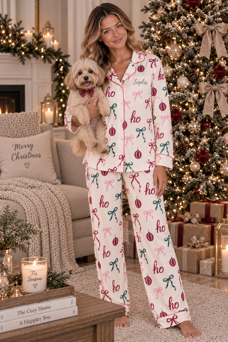A woman wearing a cozy Christmas-themed pajama set stands in a decorated living room, holding a small fluffy dog. The pajamas are cream-colored with a repeating pattern of red ornaments and green bows, with the word 'holidays' printed on the chest.