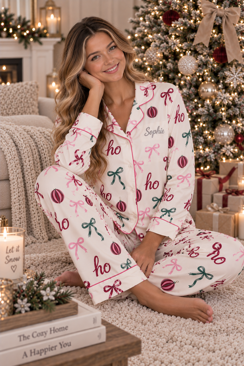 A woman sits on a fluffy cream rug in a cozy living room, wearing a pajama set with a holiday print. The pajamas are cream-colored with pink and green bow and ornament motifs, and the name 'Sophie' is embroidered on the chest.
